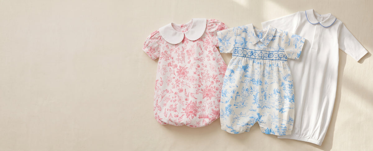 Flat lay composition of three luxury heirloom baby outfits—a pink floral bubble dress, a blue toile smocked romper, and a white pima gown with blue piped trim—arranged on a soft ivory textured background with open space on the left for text.