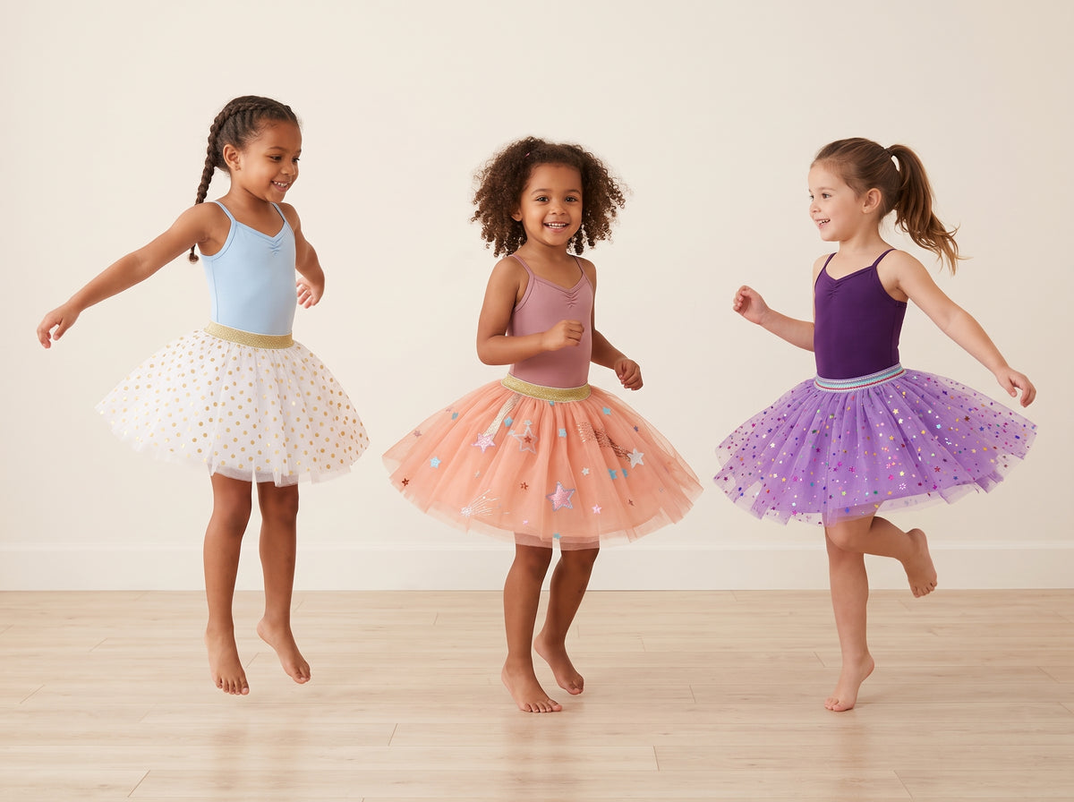 Three young girls wearing colorful tutus and leotards are jumping together in a bright studio with a light beige wall and wood floor.