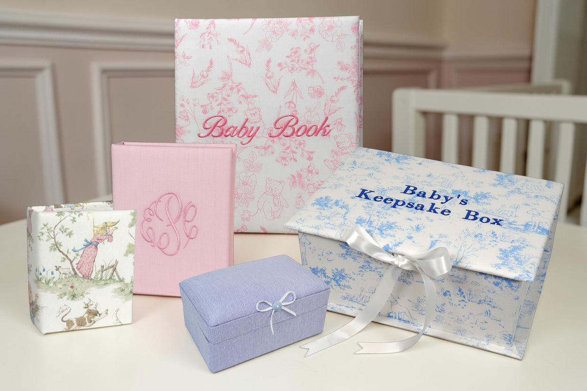 A coordinated baby keepsake gift set arranged on a tabletop, including a tall white baby book with pink floral print and pink script text, a pink album, a lavender jewelry box with a white bow, and a blue-and-white floral baby keepsake box tied with a white ribbon. A small wrapped item with a whimsical illustration stands on the left.
