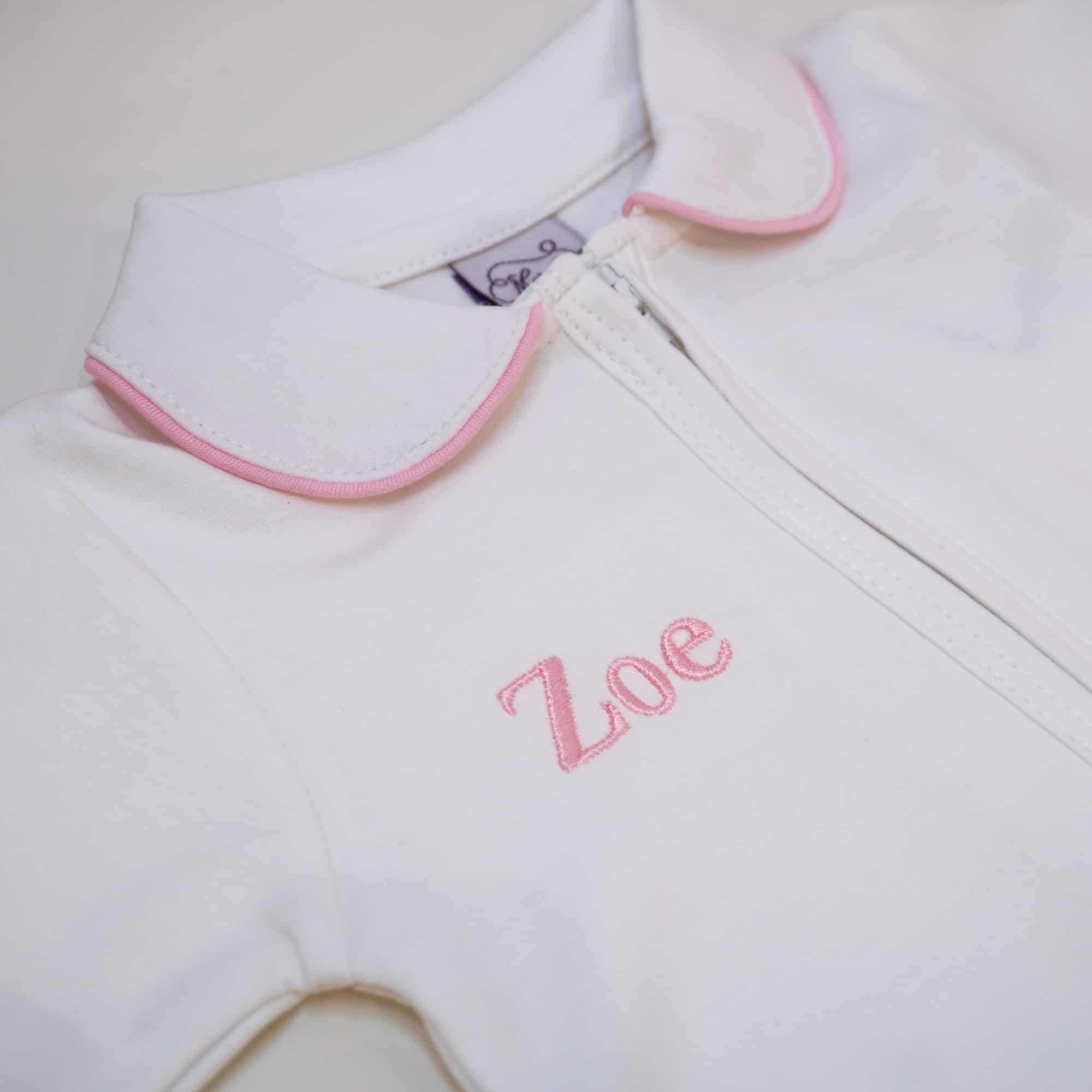 White Pima cotton baby footie with pink-trimmed Peter Pan collar and front zipper, embroidered with the name ‘Zoe’ in bodoni font and light pink thread.